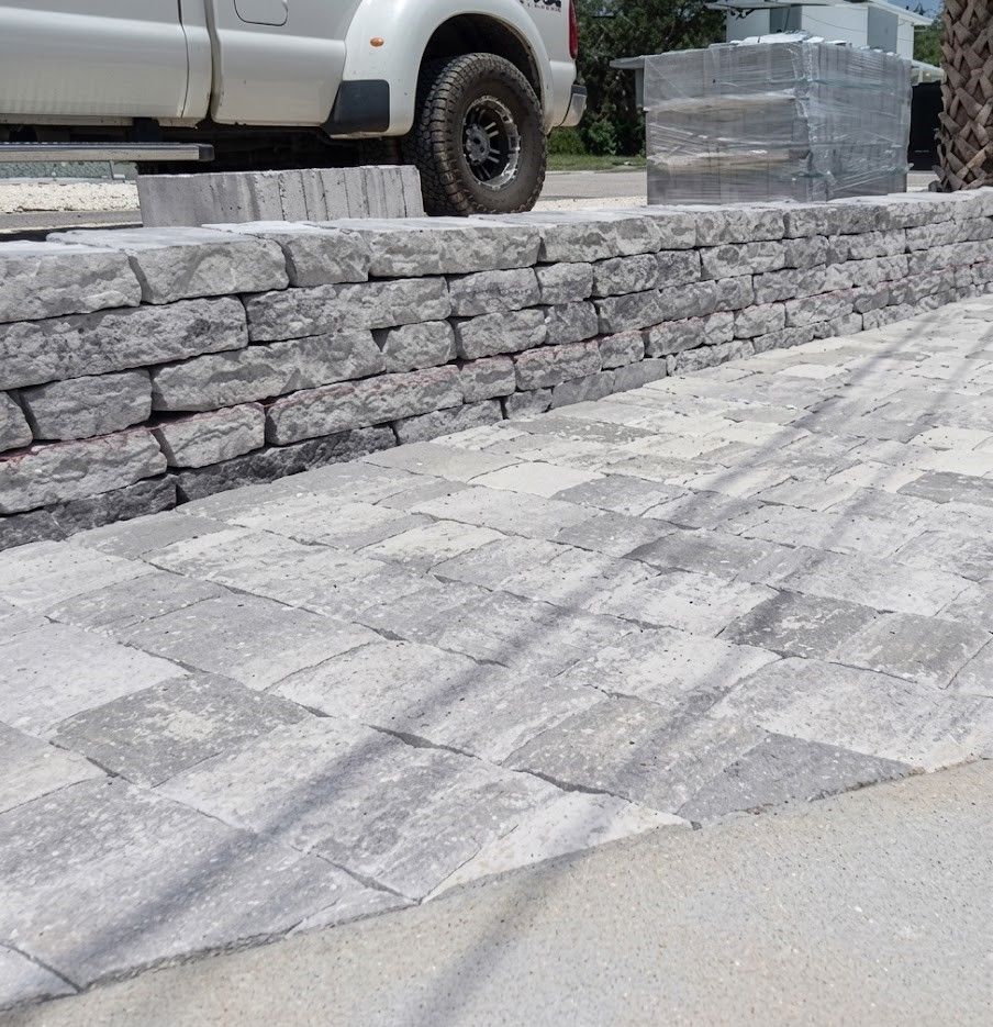 Workers laying grey paving stones on a prepared gravel base, with a wheelbarrow in the background.