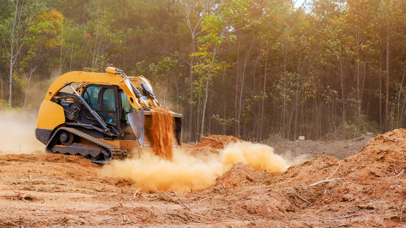 Yellow skid steer, with tracks, moving dirt in a clearing with trees in the background.