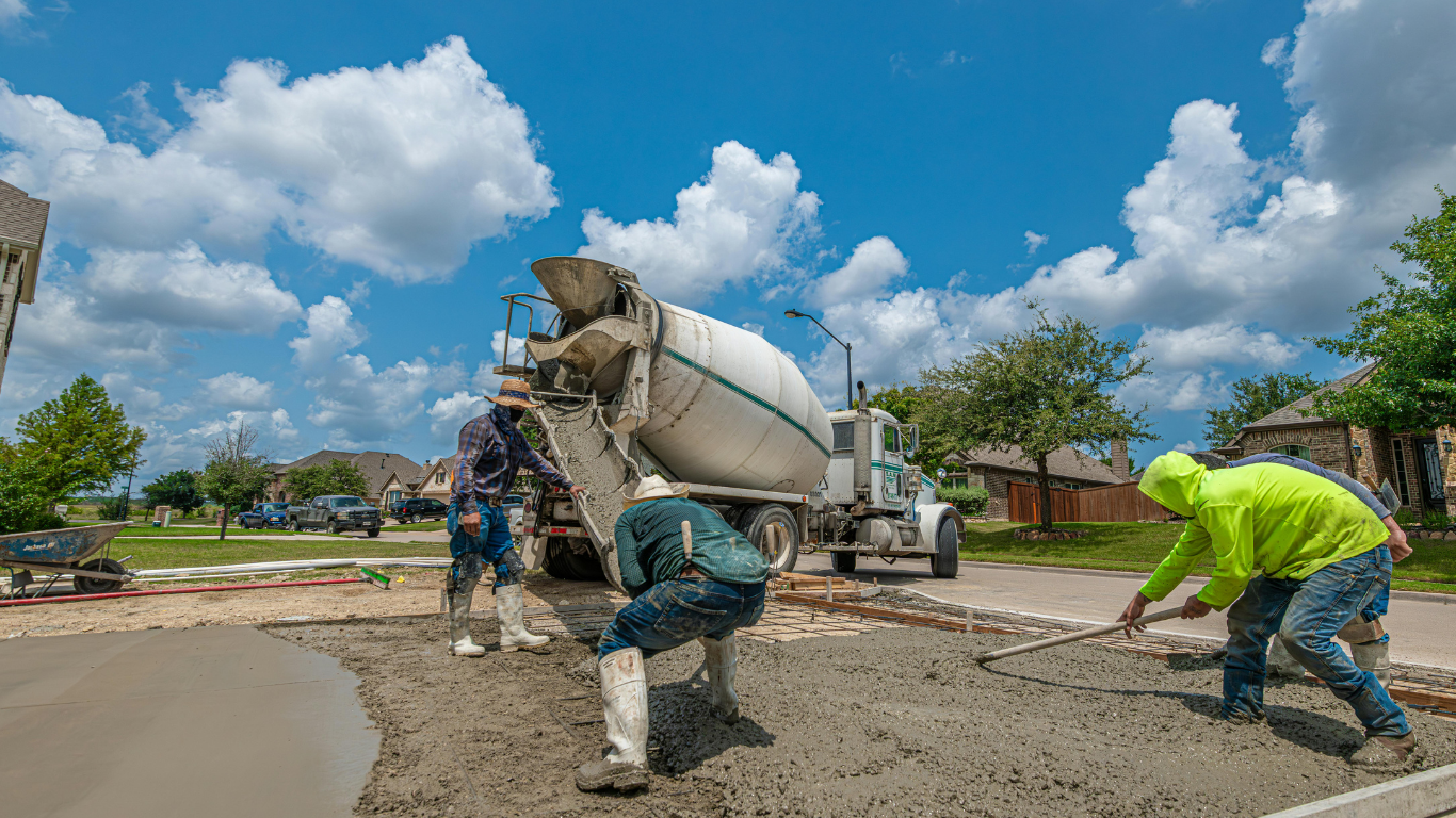 Concrete being poured from a truck onto a road by construction workers under a blue sky.