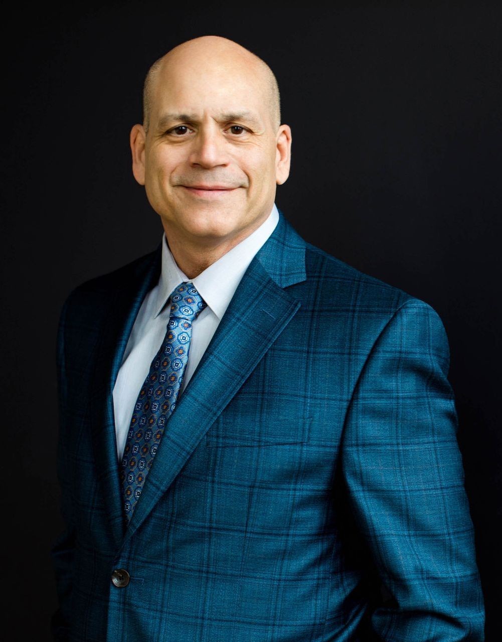 A bald man in a blue suit and tie is standing in front of a black background.