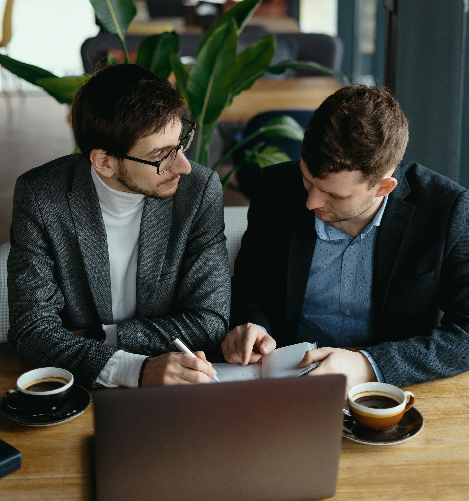 Two men are sitting at a table with a laptop and cups of coffee.