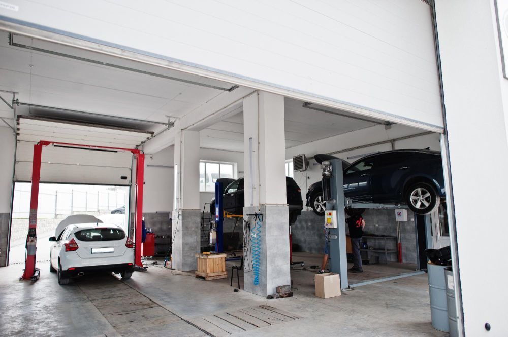 Inside a car repair shop, two cars are present: one raised on a lift, one on the ground with its hood open. Inside a car repair shop, two cars are present: one raised on a lift, one on the ground with its hood open.