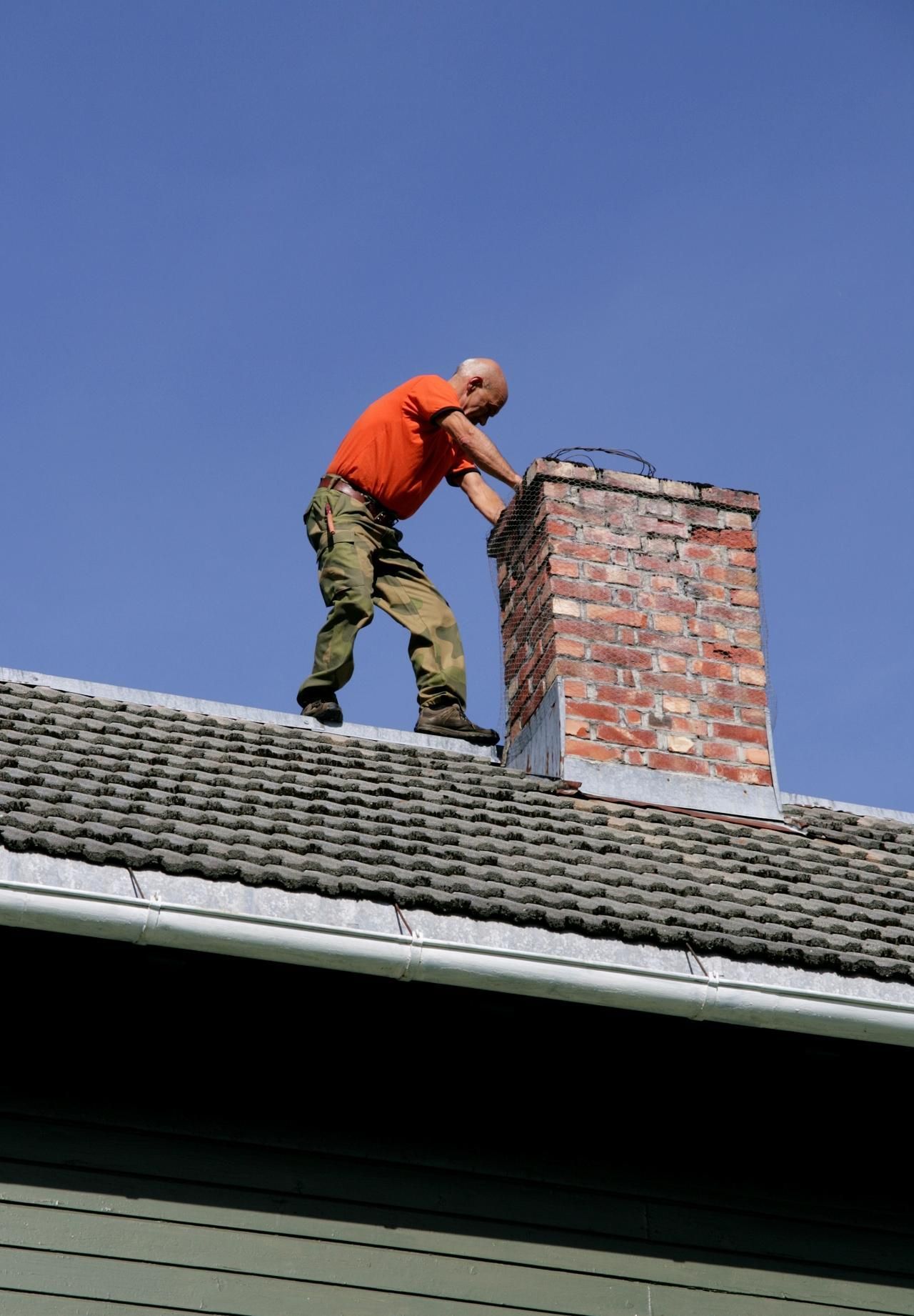 A man in an orange shirt is working on a chimney on the roof of a house.