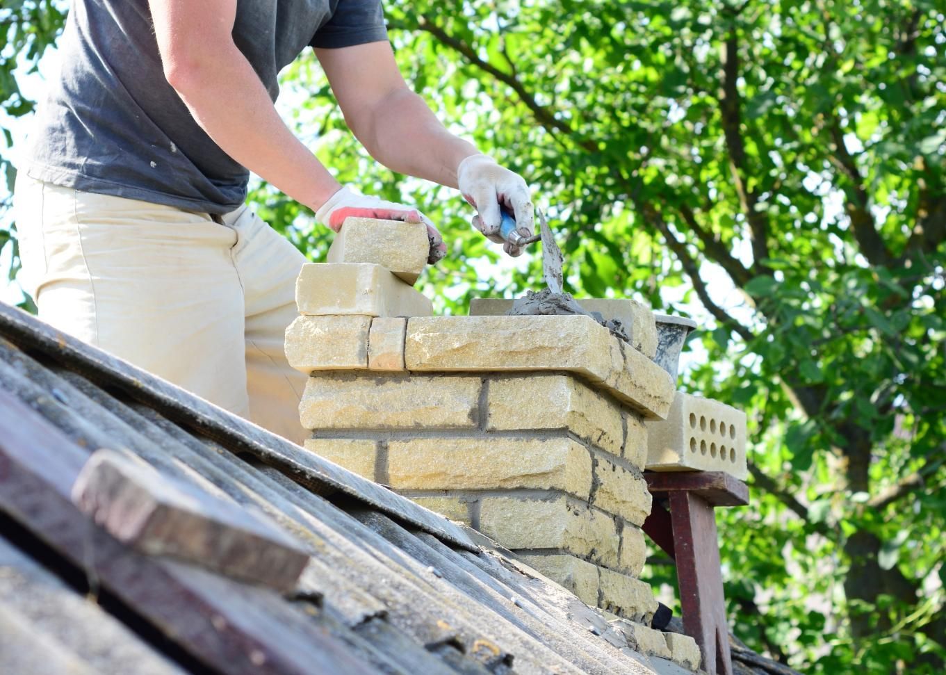 A man is laying bricks on top of a chimney on a roof.