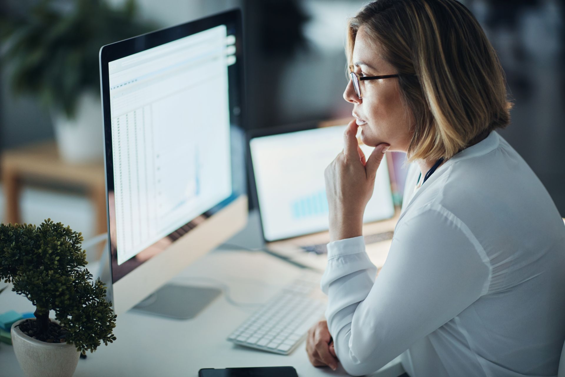 Woman with glasses looks at a computer screen, hand on chin, in an office.