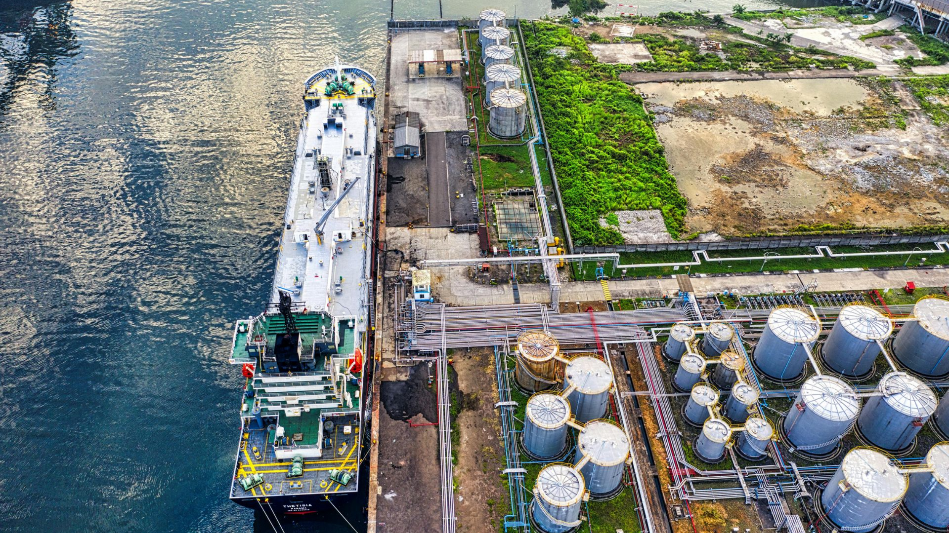 Oil tanker docked at a port next to storage tanks and industrial infrastructure.