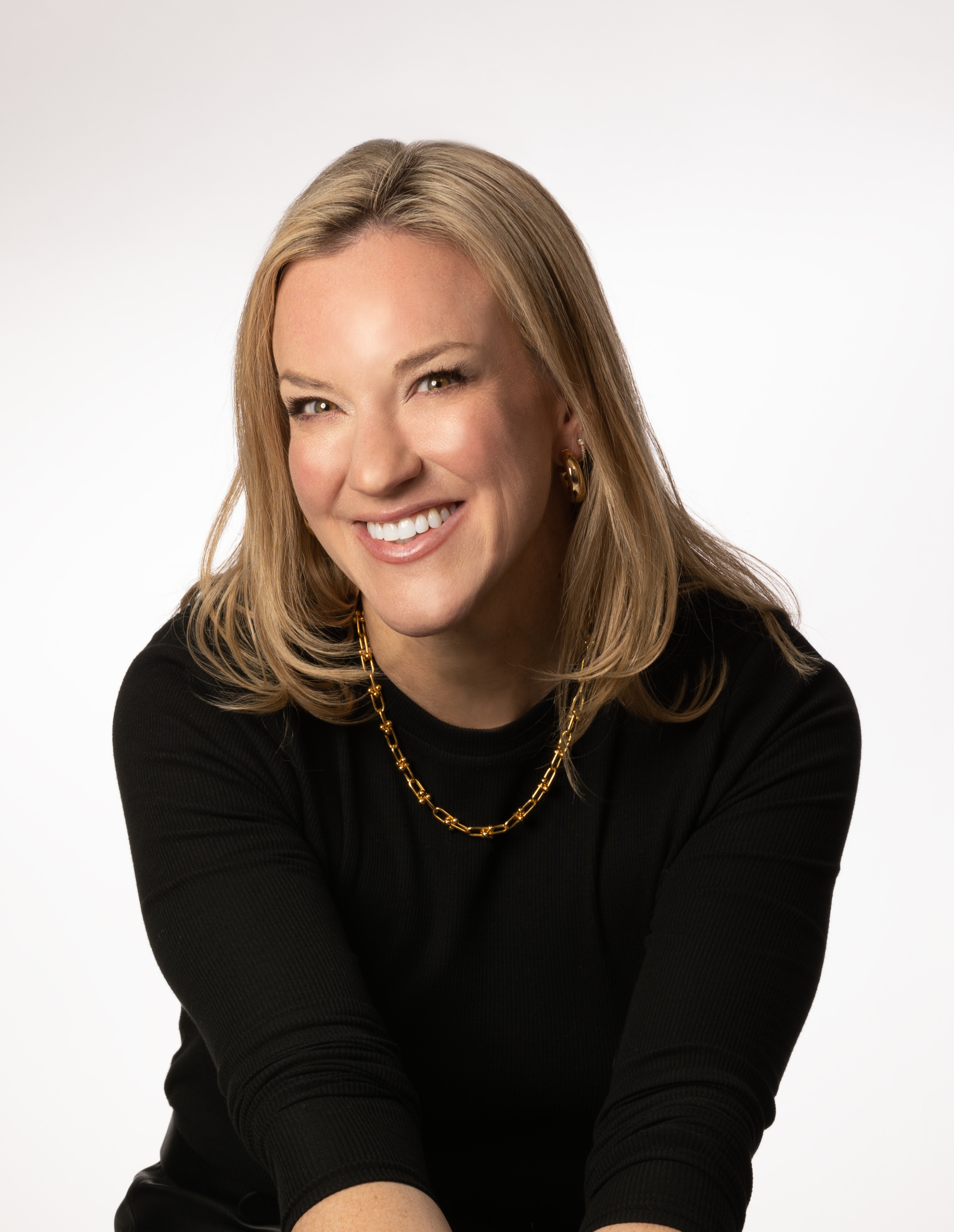 MBO Media Founder, Mary Beth Olson smiling in a black top and gold necklace, leaning forward against a white background