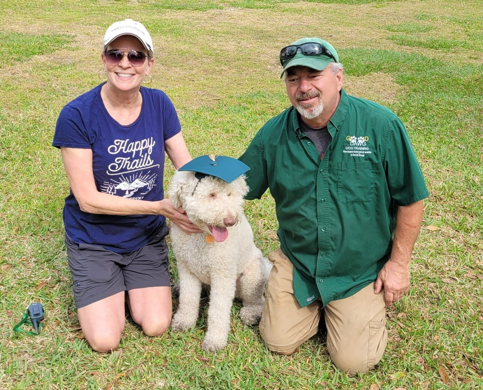 A man and a woman are kneeling next to a dog wearing a graduation cap.