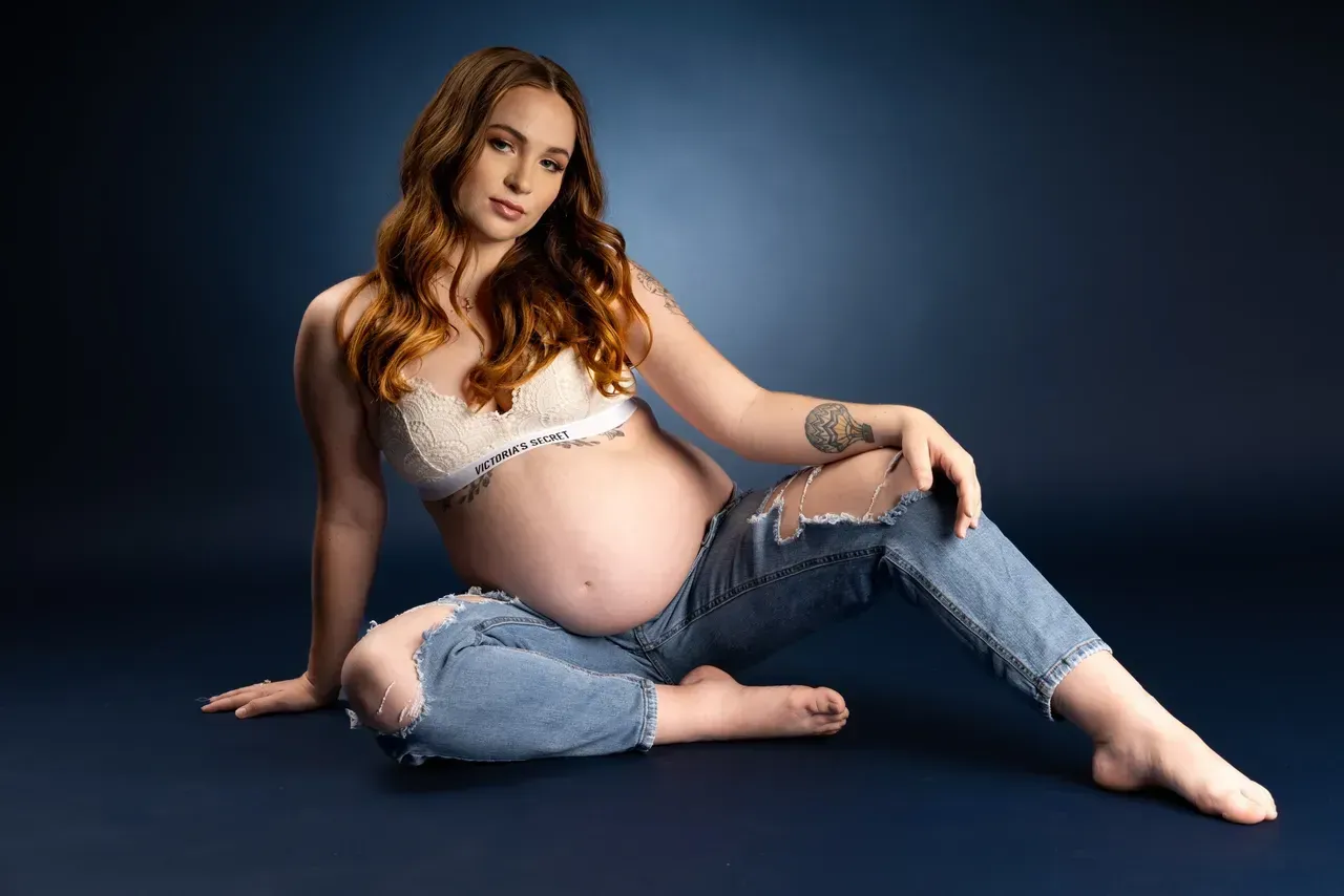 A pregnant woman is sitting on the floor wearing jeans and a bra.