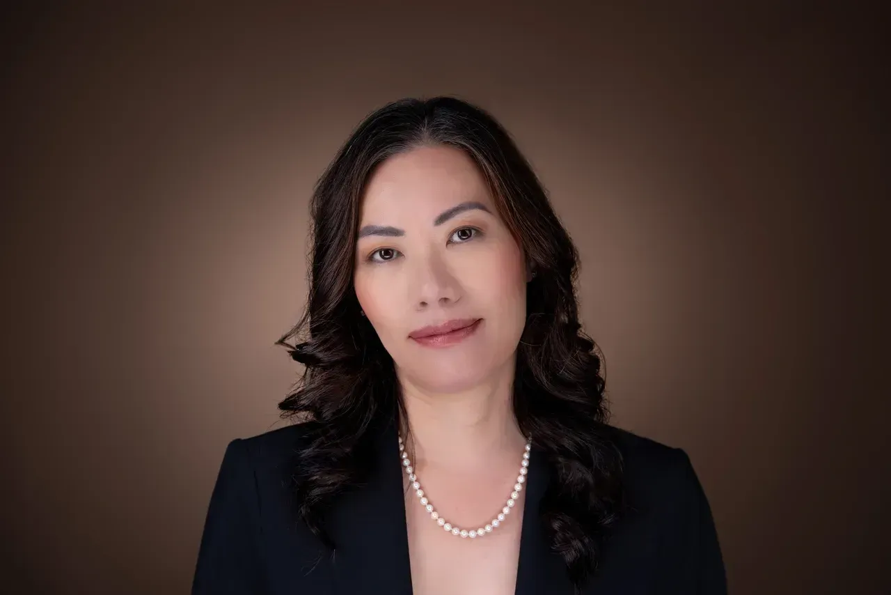 A woman wearing a pearl necklace and a black jacket is standing in front of a brown background.