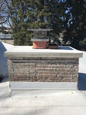 Brick chimney with a metal cap on a flat roof, against a backdrop of trees and a blue sky.
