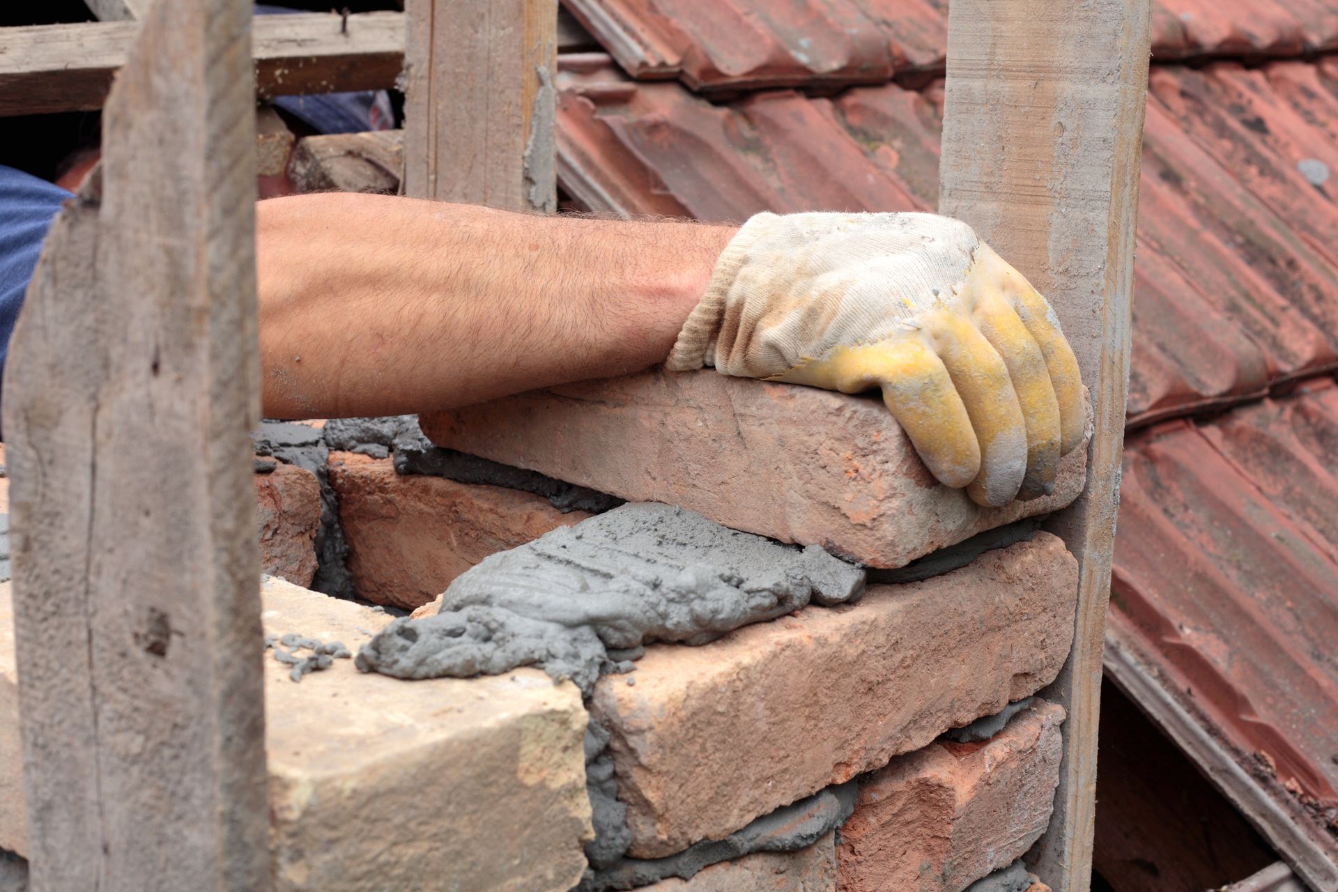 A bricklayer, wearing a glove, is laying bricks with mortar on a rooftop, with wood framing around the bricks.