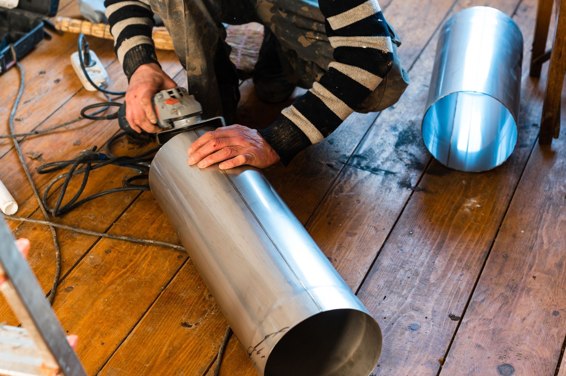Person using a grinder to cut a metal cylinder on a wooden floor, another cylinder nearby.