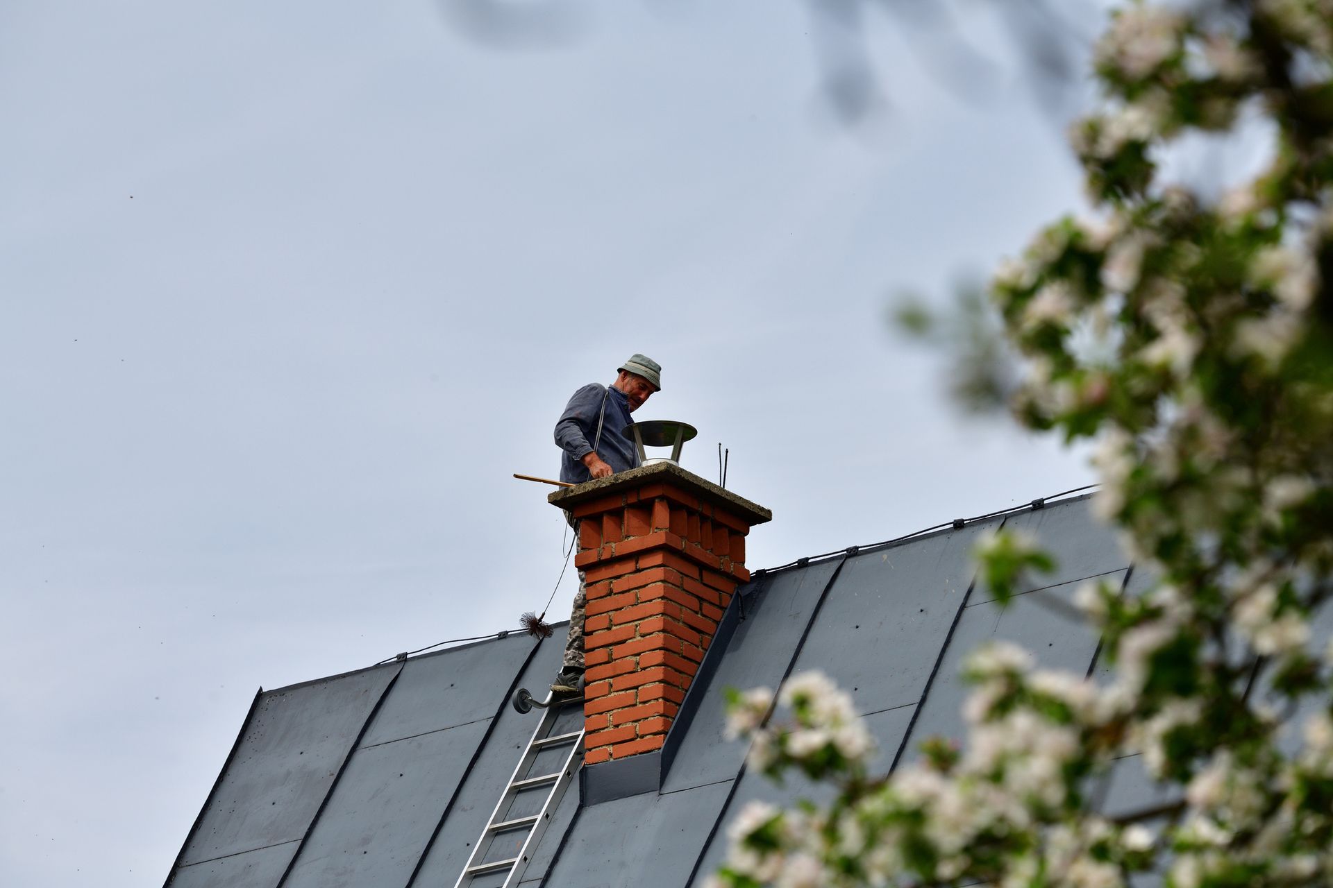 A person on a roof inspecting a brick chimney. A ladder is propped against the roof.