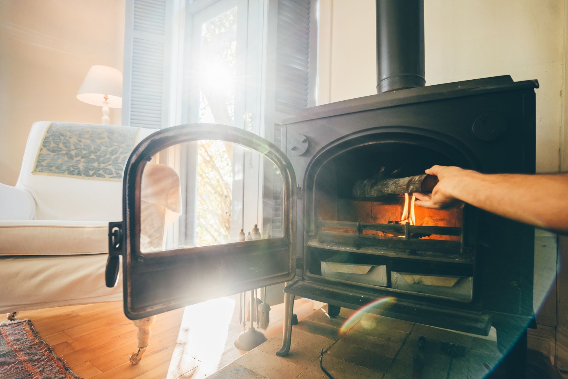 Hand placing wood in a lit, black wood-burning stove in a warm, sunny room.