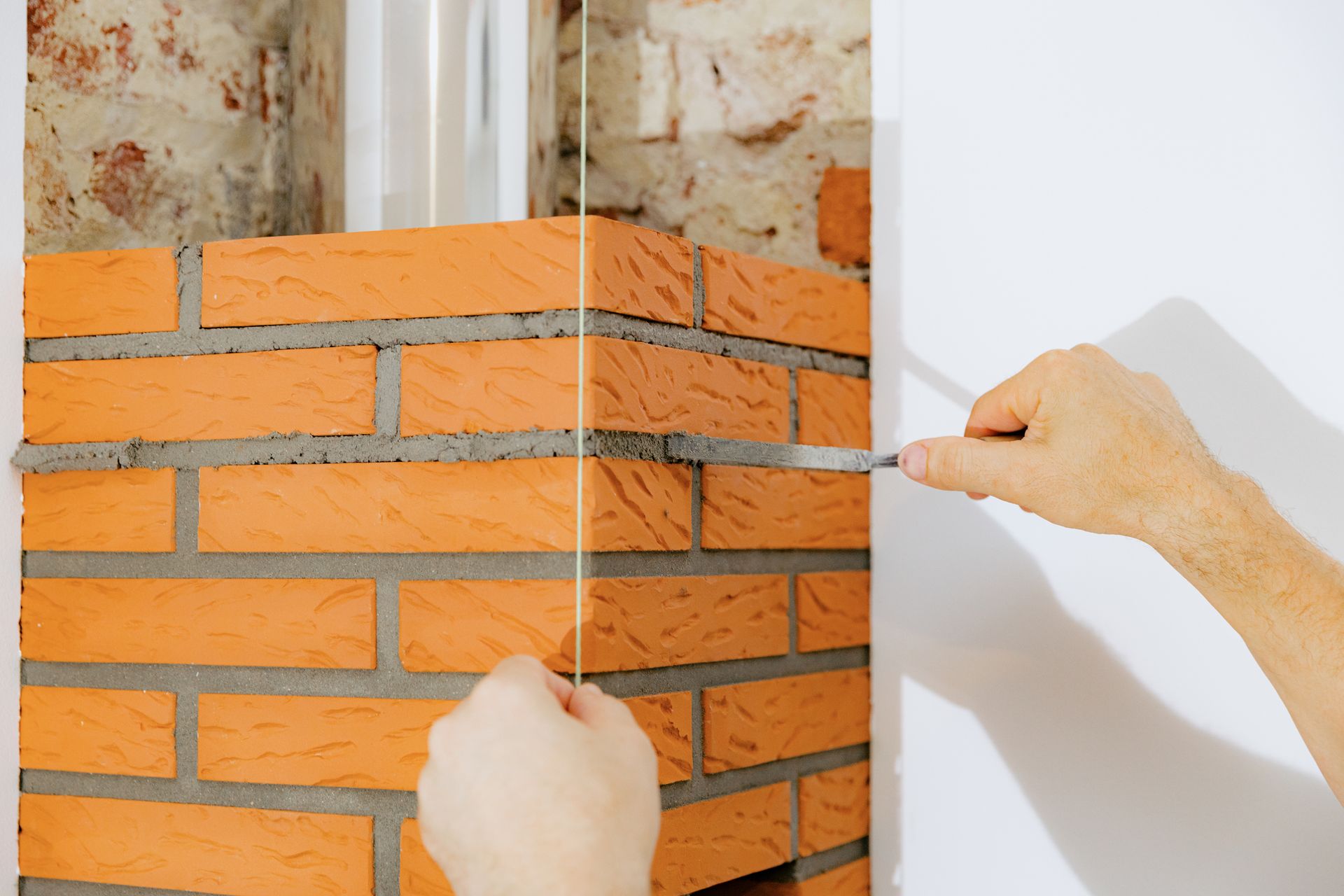 Hands are shown using a string and trowel to check a corner brick wall being built indoors.