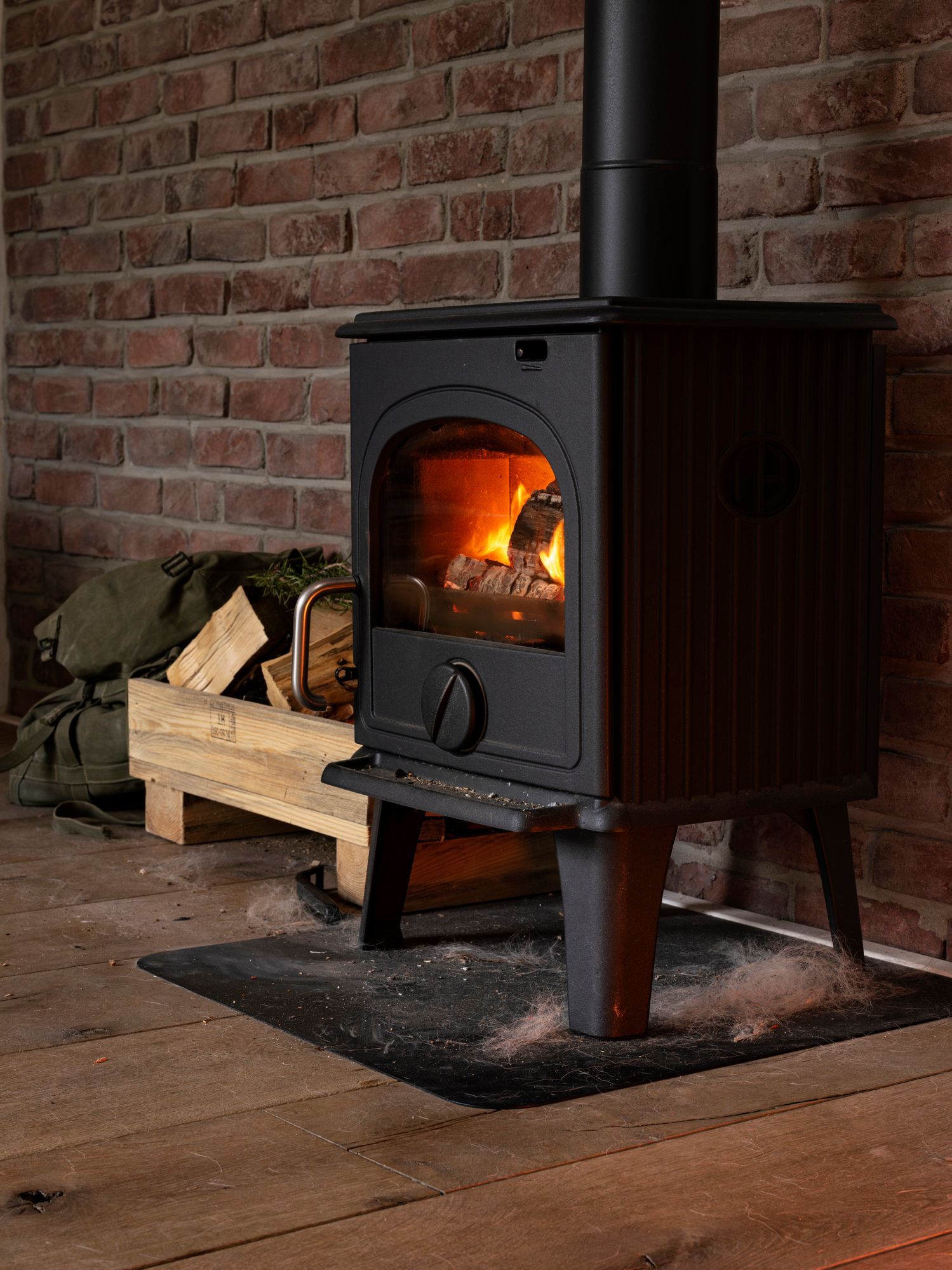 Wood-burning stove with flames, in front of a brick wall and wooden floor. Wood pile nearby.