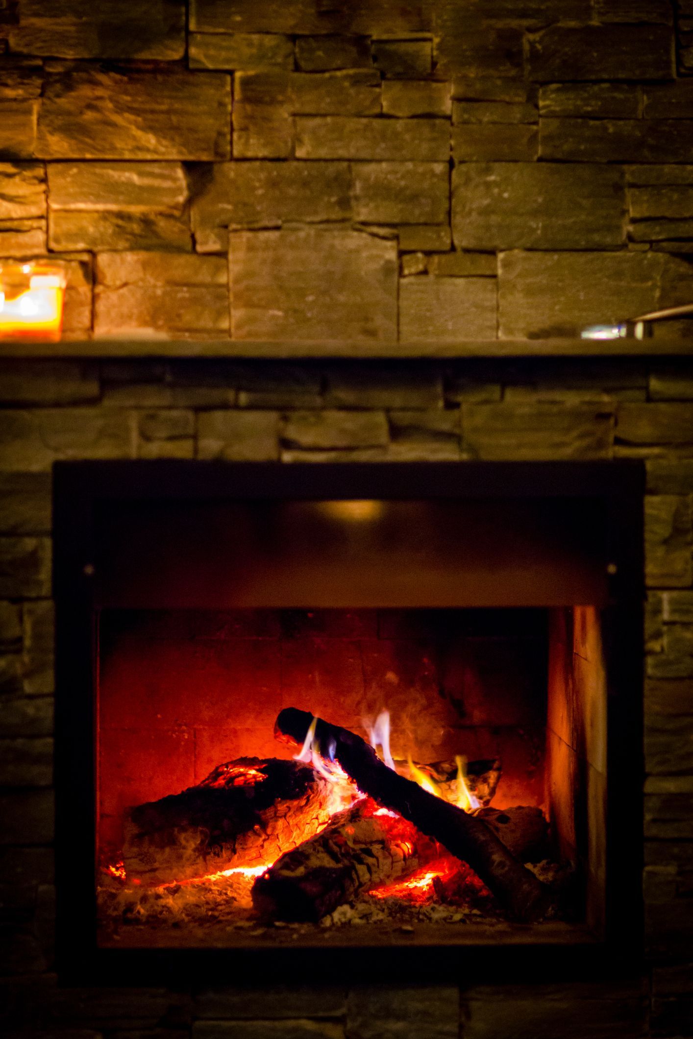 Fireplace with glowing embers and logs against a stone wall, lit by soft orange light.