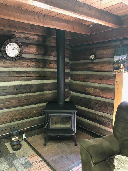 Wood-burning stove in a log cabin corner, black, with chimney. A clock hangs on the log wall, and a green chair is visible.