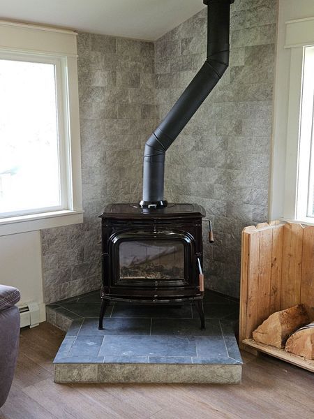 Wood-burning stove in corner, flanked by windows. Grey tiled wall and hearth, black stovepipe.