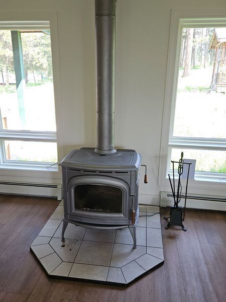 Gray wood-burning stove with chimney in a room, flanked by windows. A fire tools stand is to the right.