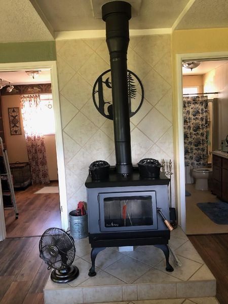 Wood-burning stove with black pipe, in a room with light-colored tile and a black fan.