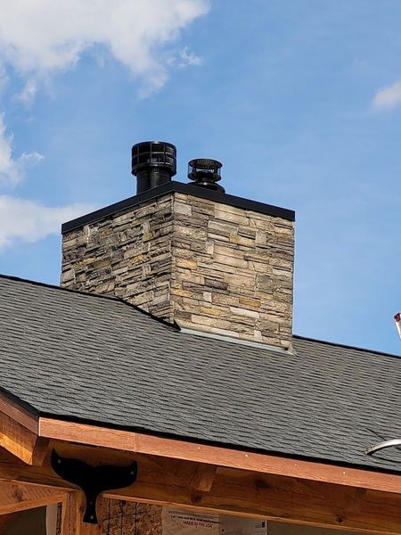 Stone chimney with black caps on a dark shingled roof under a blue sky.