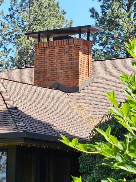 Brick chimney on a brown shingled roof, topped with a black cap, set against a blue sky.