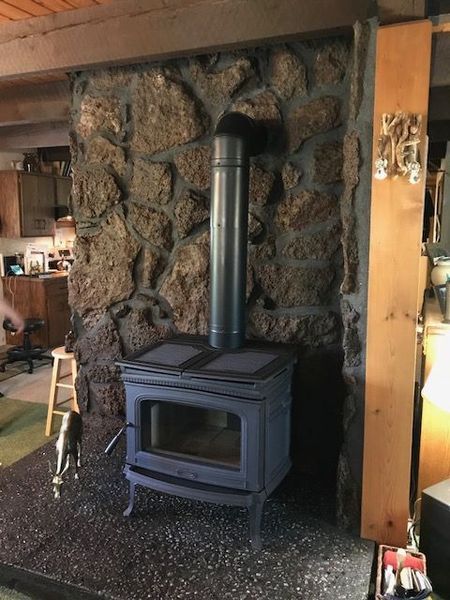Wood-burning stove with metal flue pipe against stone wall, with a small pet and light in the foreground.