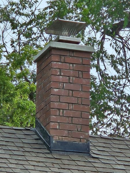 Brick chimney with a metal cap on a shingled roof, trees in background.