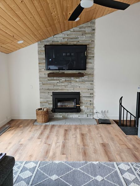 Living room with stone fireplace, TV, and wooden ceiling. Hardwood floors and area rug.