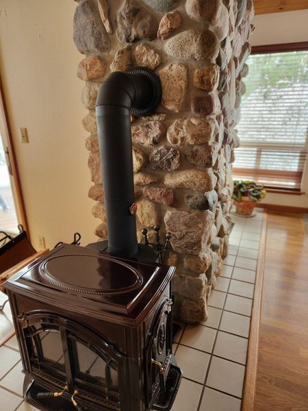 Dark brown wood stove with black pipe against a stone chimney, on a tiled hearth.