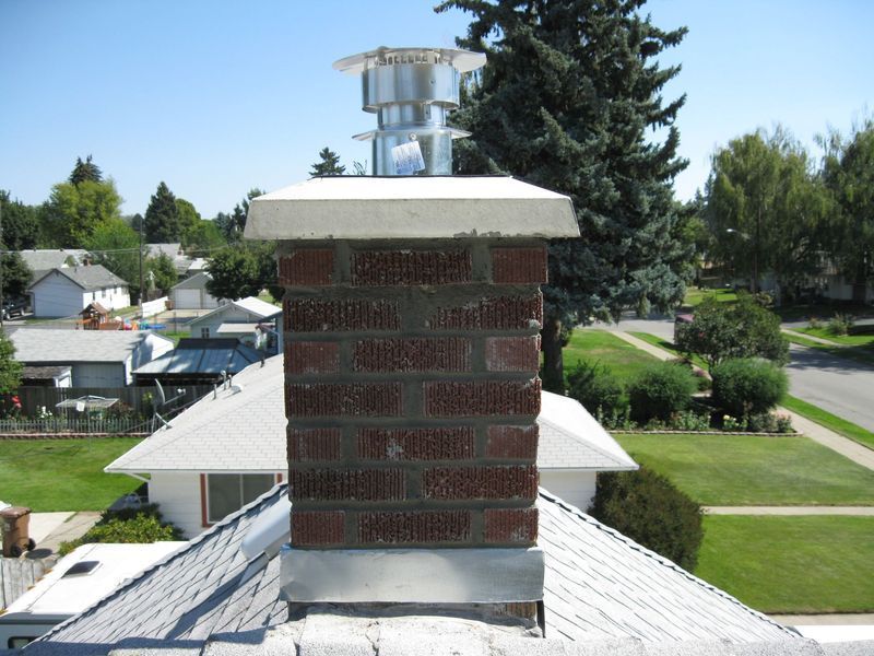 Brick chimney with metal cap on a residential roof, with a neighborhood in the background.