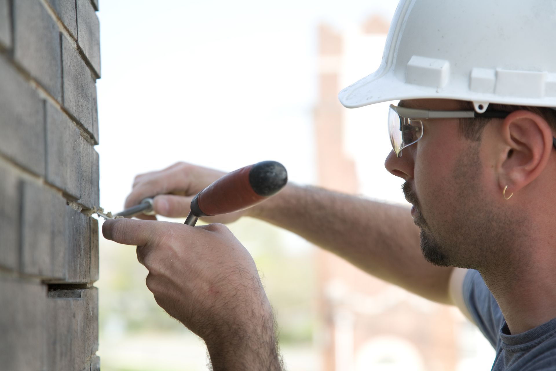 Construction worker using a trowel to work on brickwork, outdoors. Wearing a white hard hat and sunglasses.