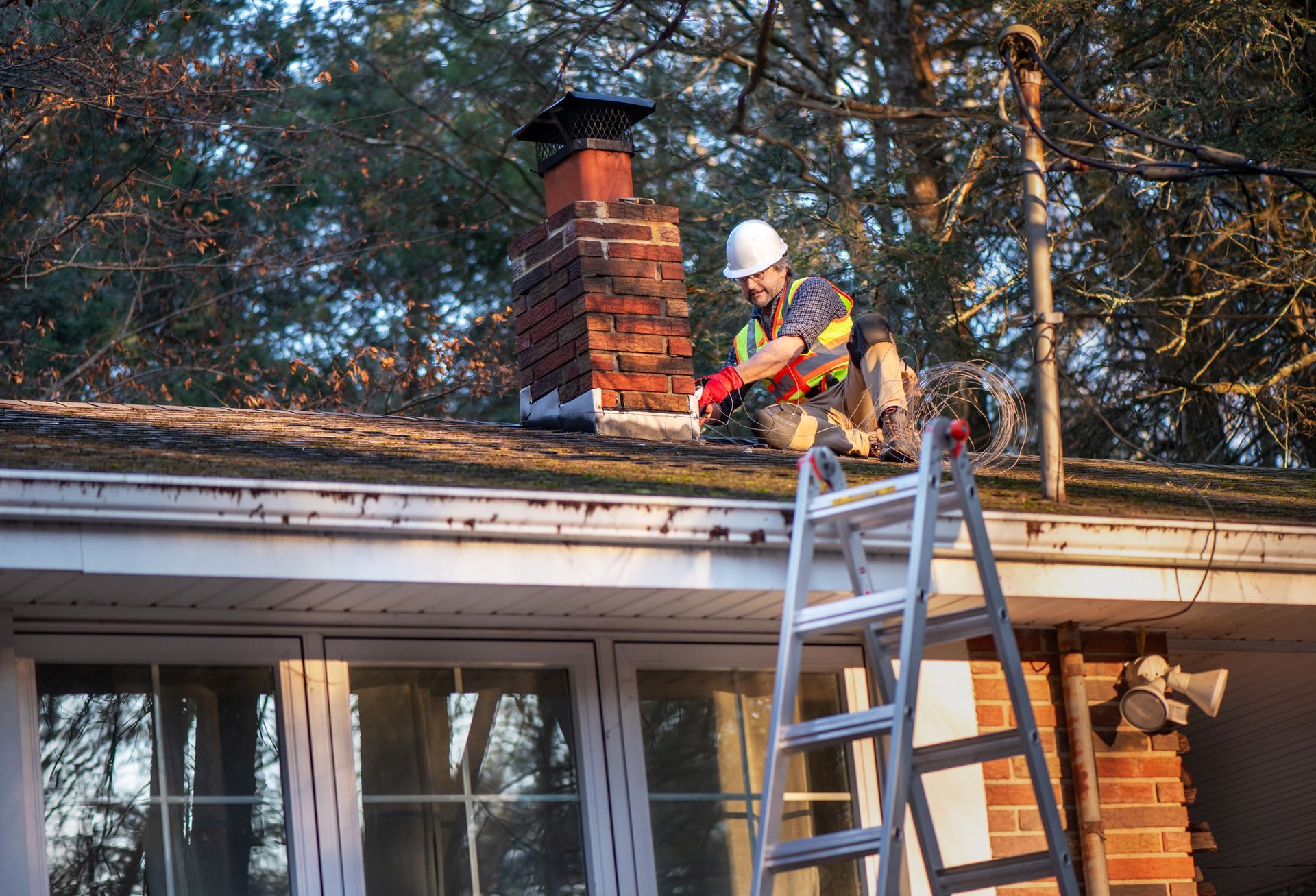 Roofer in safety gear on a roof near a brick chimney. Ladder visible.