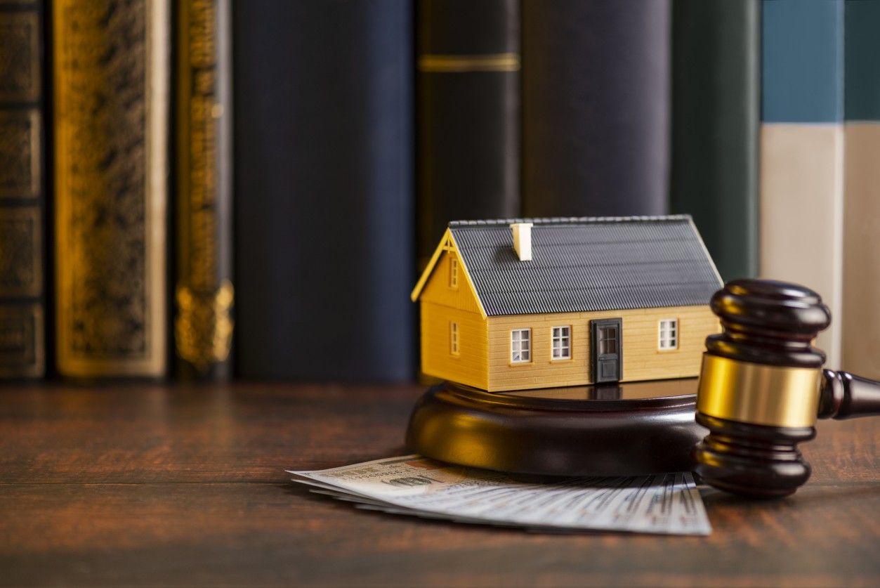 A house model and a judge's gavel rest on a wooden surface surrounded by books and money