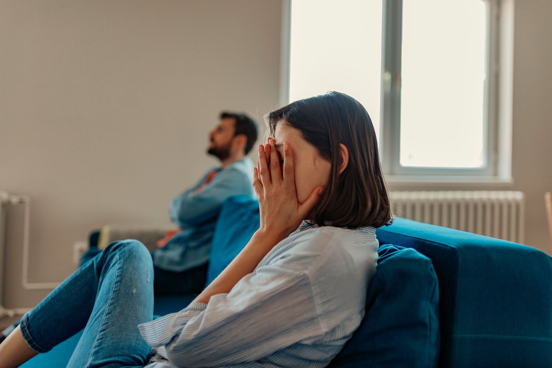 A woman sits on a sofa, her hands covering her face, expressing a sense of distress