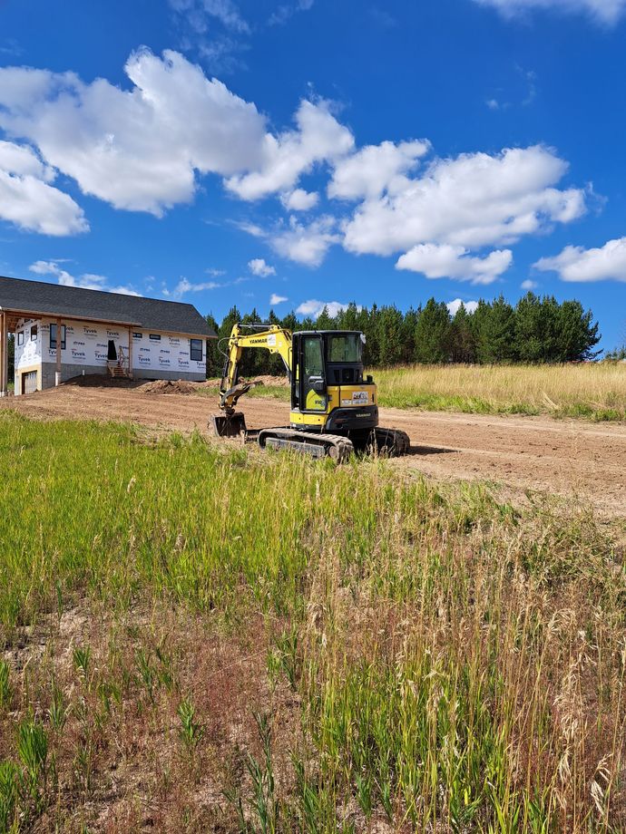 A yellow excavator is driving through a grassy field in front of a house under construction.