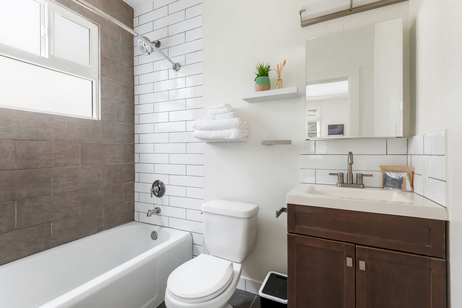 Bathroom with white and gray tile, tub, toilet, vanity, and small shelf with decor.