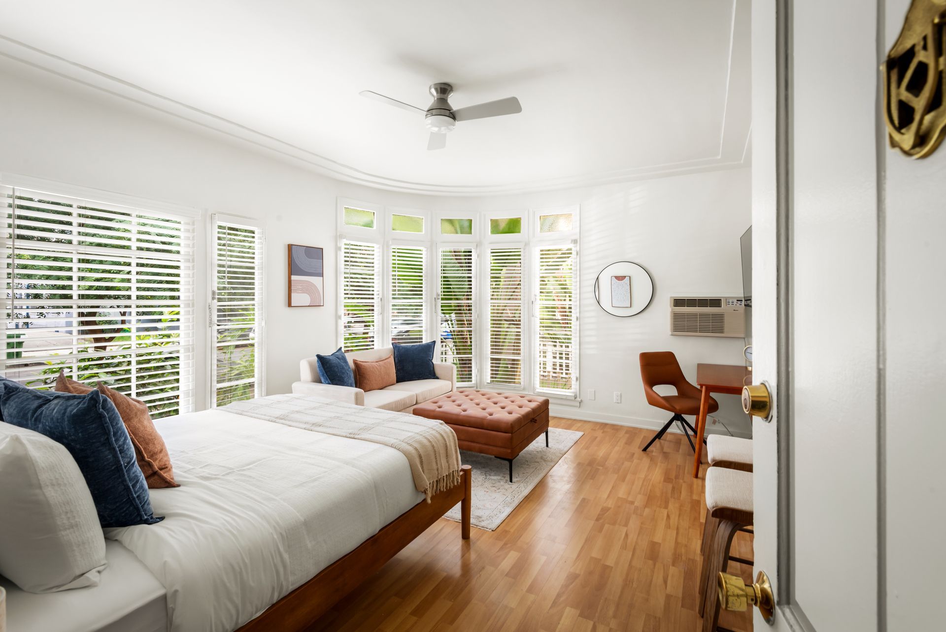 Bright studio apartment with white walls, wood floors, and large windows. Furnished with a bed, sofa, and desk.