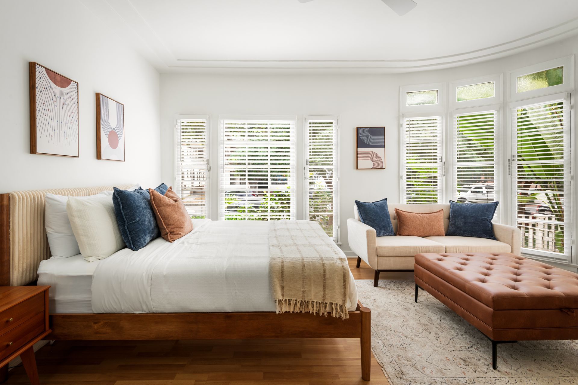 Bedroom with a bed, small sofa, and a leather ottoman in front of a window with shutters; light, bright space.
