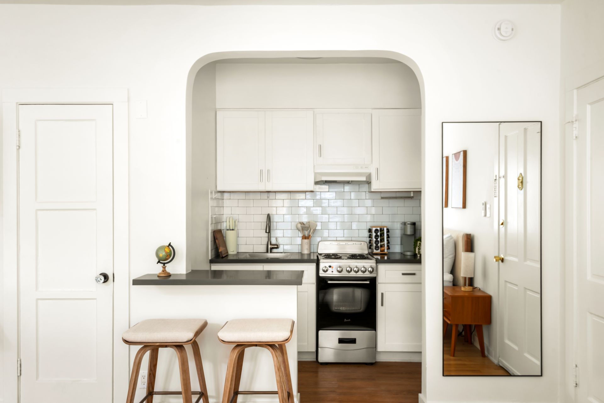 Small kitchen with white cabinets and a dark countertop, separated by an arched doorway. Two stools sit at a breakfast bar.