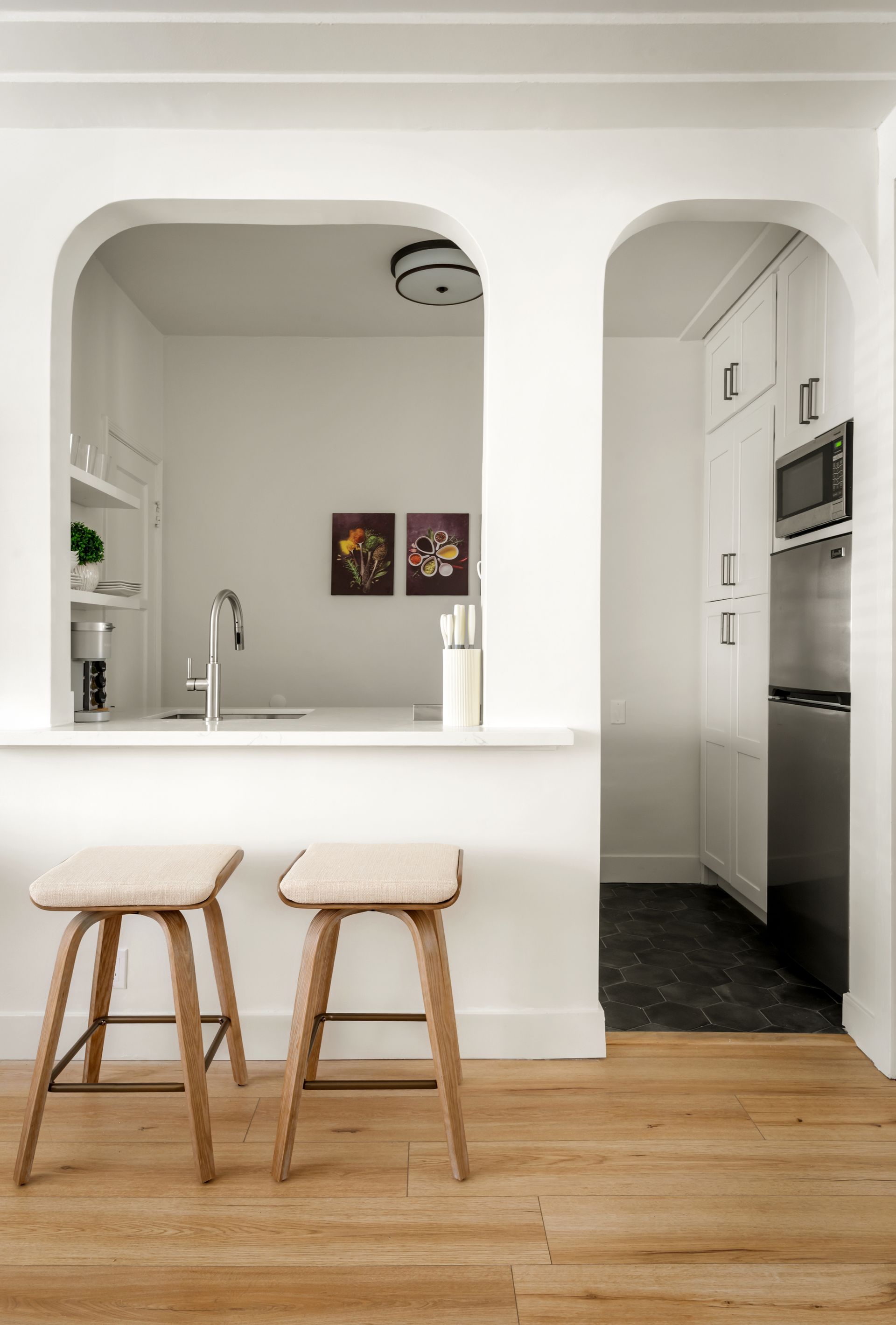 Two stools at a kitchen pass-through; white walls, wood floors, stainless steel appliances.