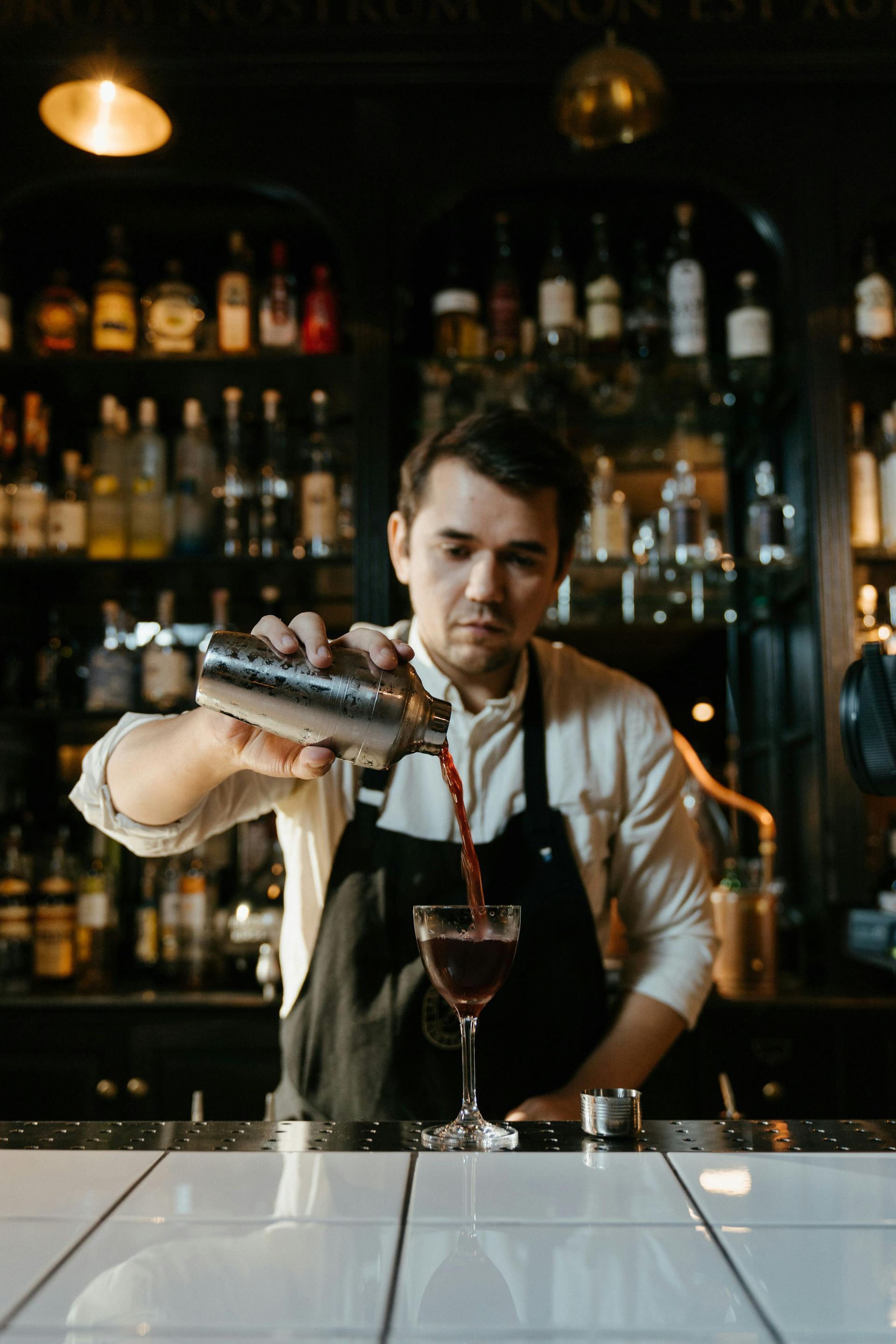 Bartender pouring cocktail from shaker into a glass, behind a bar with liquor bottles.
