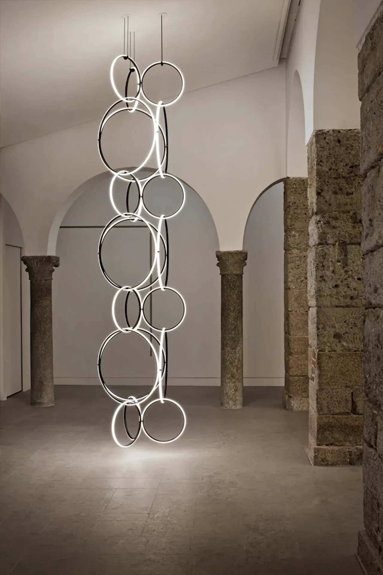 Lampadario moderno con anelli circolari luminosi, appeso in una stanza con colonne in pietra.