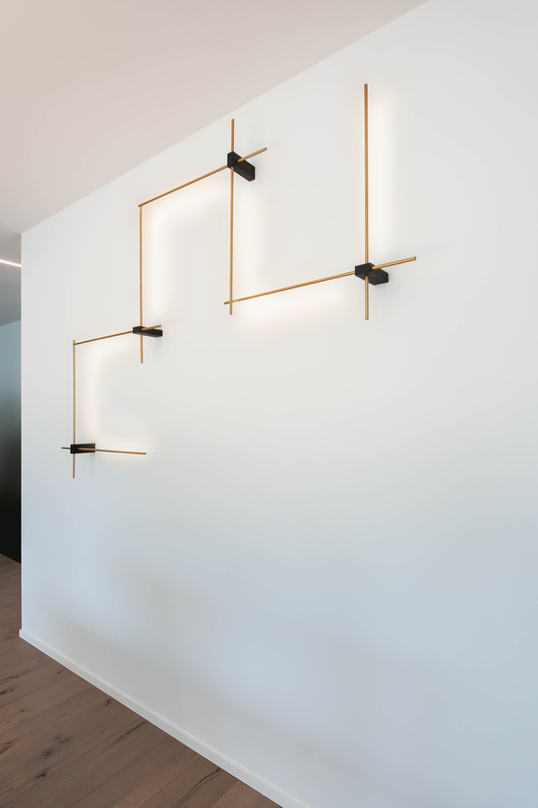 Applique lineari dorate con giunti neri su una parete bianca, sopra un pavimento in legno.