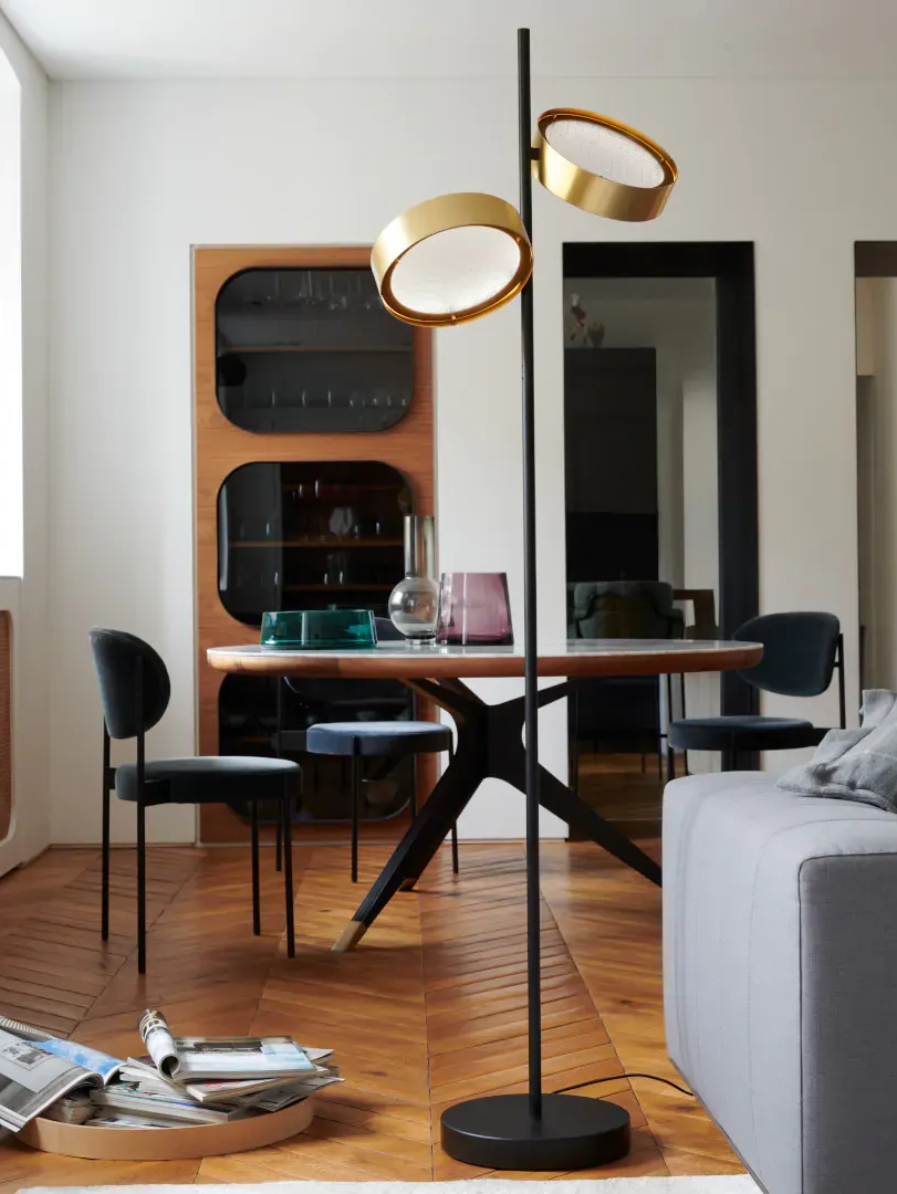 Lampada da terra con due paralumi dorati a forma di disco in una moderna sala da pranzo, che illumina un tavolo e delle sedie.