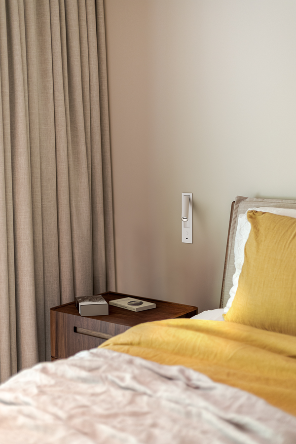 Angolo della camera da letto con comodino, letto e tenda. Biancheria da letto gialla e grigia, pareti neutre, interruttore della luce bianco.