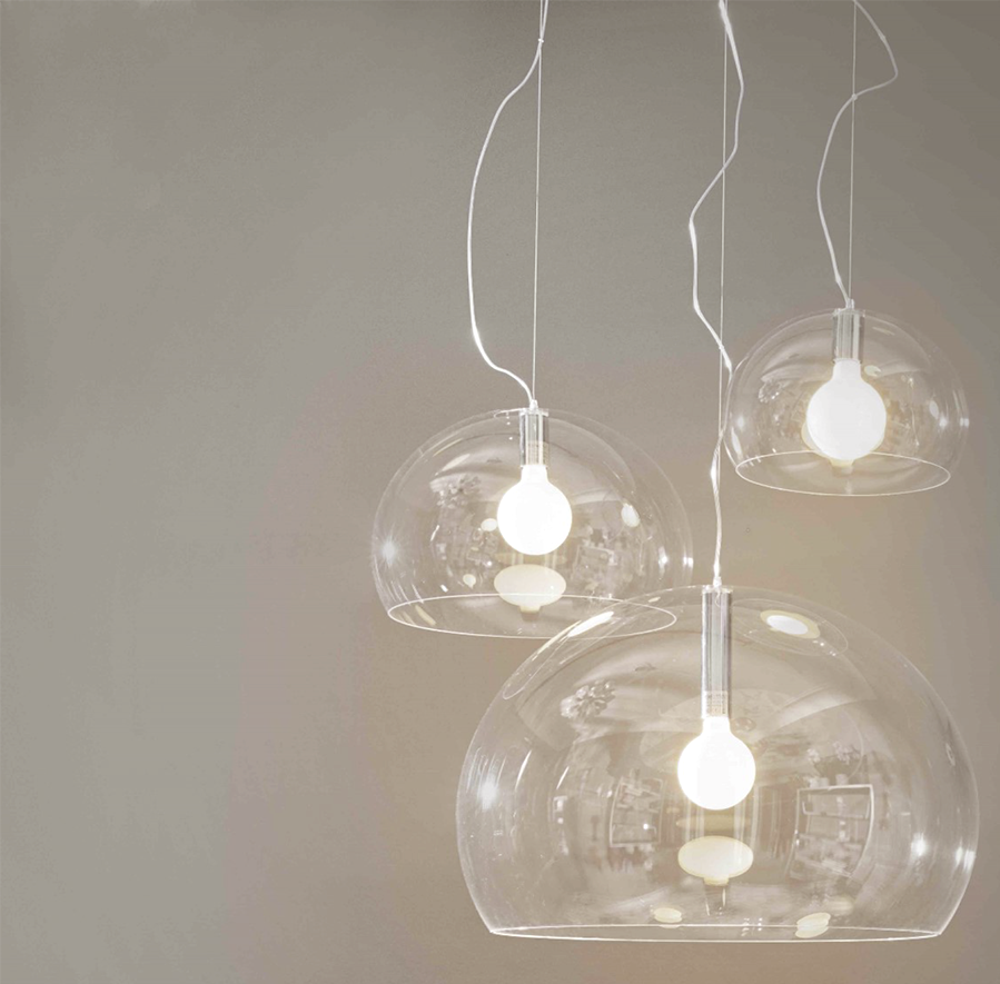 Tre lampade a sospensione a cupola in vetro trasparente con lampadine illuminate, appese a cavi bianchi contro una parete grigia.