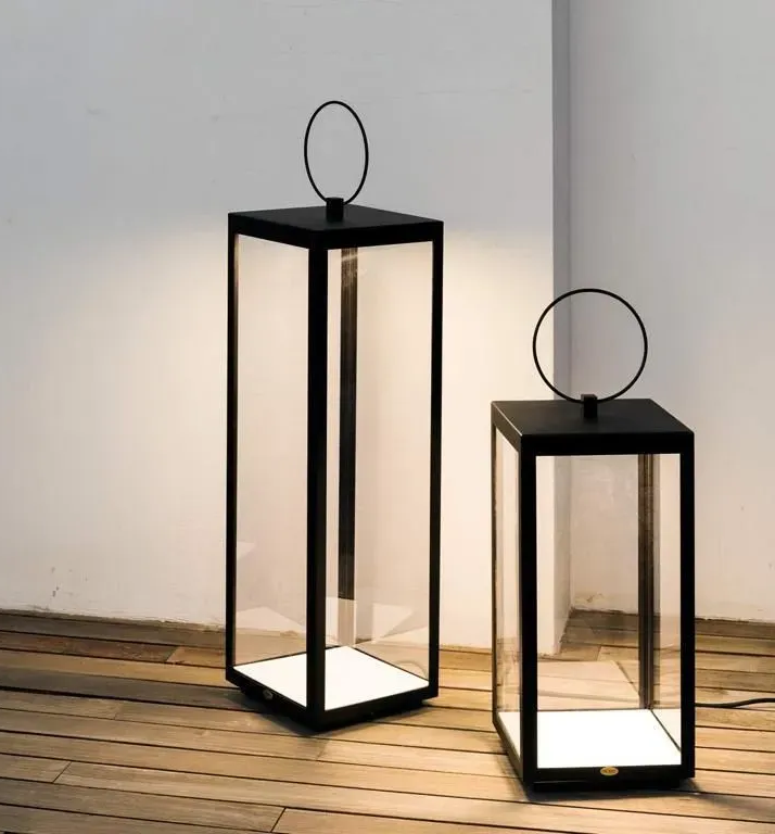 Due lanterne nere in metallo e vetro con interni illuminati, su un pavimento in legno, vicino a una parete bianca.